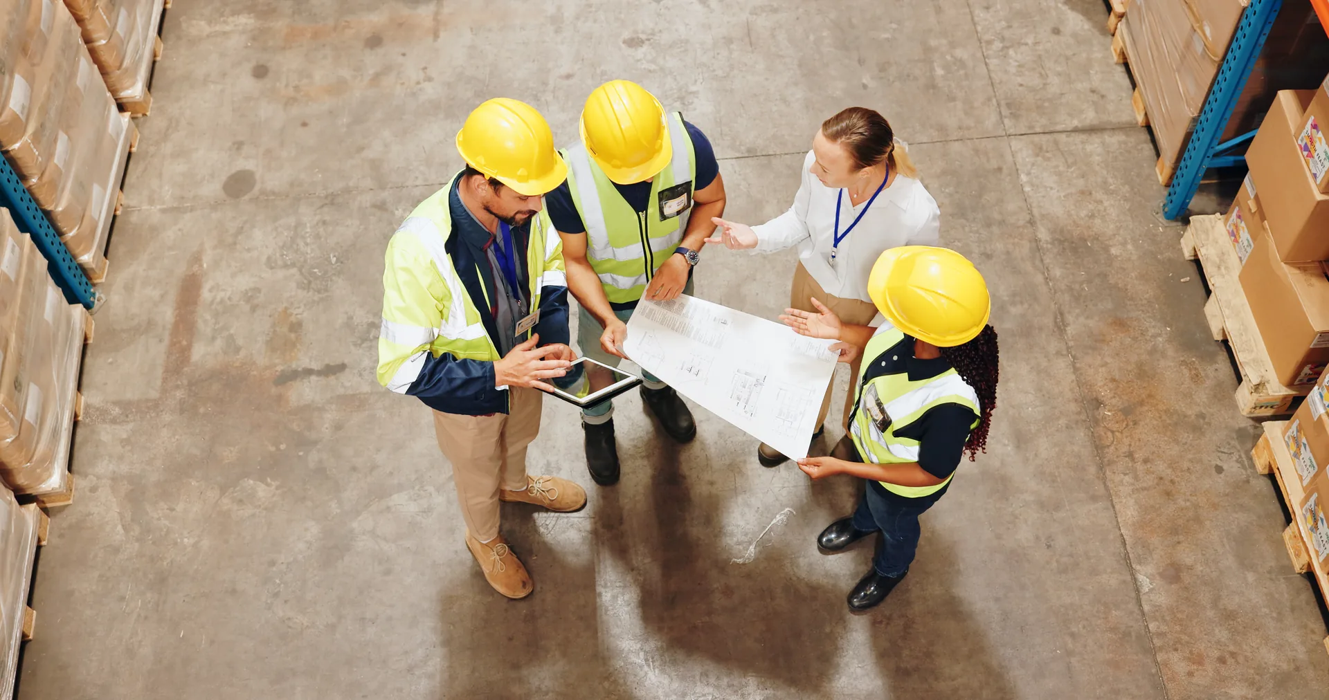 Trade team in hi-vis reviewing blueprints between warehouse storage shelves.